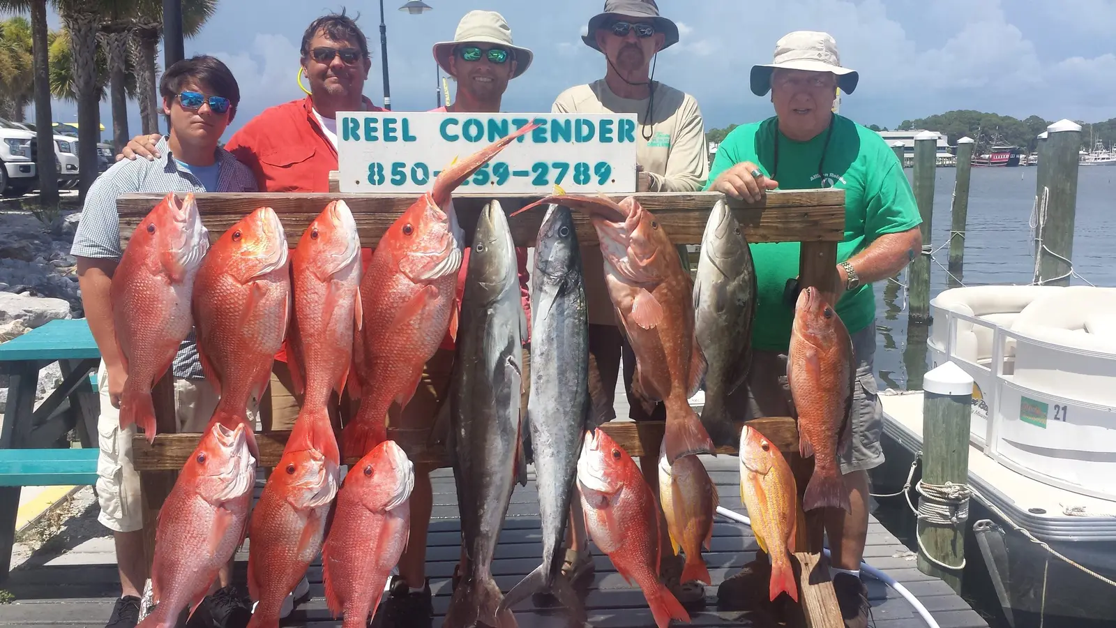 A group holding red snapper, kingfish and grouper next to the Reel Contender sign